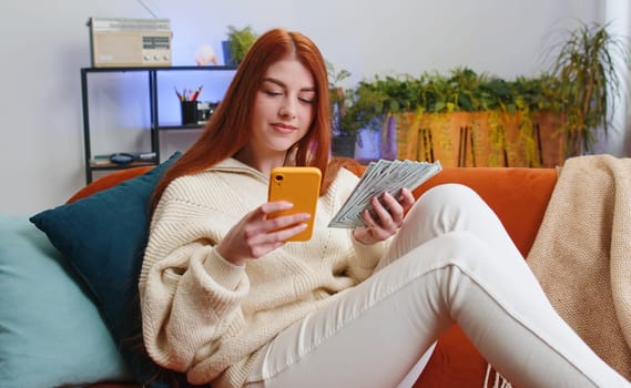 Planning family budget. Smiling redhead woman counting money cash, use smartphone calculate domestic bills at home room. Joyful girl satisfied of income and saves money for planned vacation, gifts