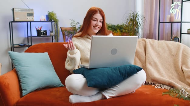 Young woman sitting on couch looking at camera, making video webcam conference call with friends or family, enjoying pleasant conversation. Portrait of redhead girl laughing waving hello at home room