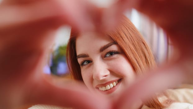 I love you. Happy redhead woman at home living room couch makes symbol of love, showing heart sign to camera, express romantic feelings express sincere positive feelings. Charity, gratitude, donation