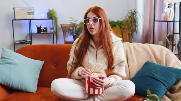 Excited young woman sitting on sofa eating popcorn and watching interesting tv serial, sport game, film, online social media movie content at home. Girl in 3D glasses enjoying domestic entertainment