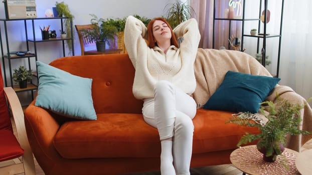 Portrait of tired young woman enjoy relaxing on home couch in room. Happy redhead girl lying on couch resting napping after hard working day, closed her eyes, put hands behind head, stress free, rest