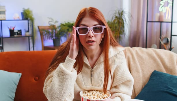 Excited young woman sitting on sofa eating popcorn and watching interesting tv serial, sport game, film, online social media movie content at home. Girl in 3D glasses enjoying domestic entertainment
