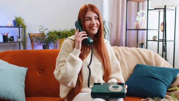 Portrait of young woman making wired telephone conversation with friends sitting call on couch at home in room. Happy excited redhead girl enjoying old-fashioned retro phone from 90s talking indoors