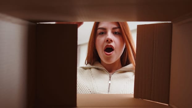 Inside view from box. Happy redhead woman unpacking delivery parcel at home. Smiling satisfied girl shopper online shop customer opening cardboard receiving purchase gift by fast postal shipping order