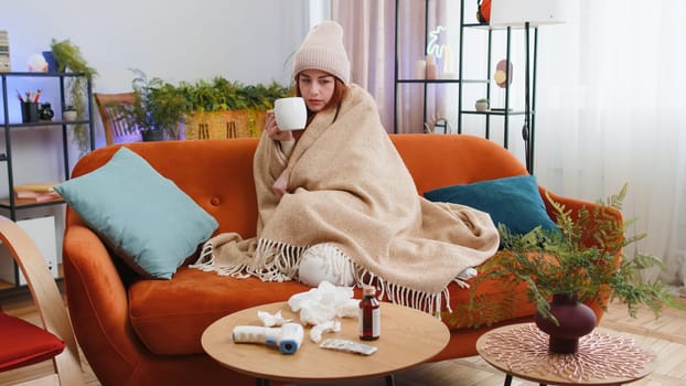 Young ill woman suffering from cold or allergy sitting on sofa in living room at home drinking hot tea. Sick redhead girl wear hat wrapped in plaid blows sneezes wipes snot into napkin. Quarantine