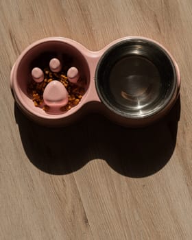 A double bowl for slow feeding and a bowl of water for the dog. Top view of a pink plate with dry food on a wooden floor