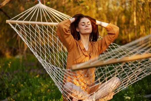 a joyful woman is sitting in a mesh hammock in nature, relaxing and enjoying the rays of the setting sun, straightening her hair with her hands, closing her eyes on a warm summer day. Horizontal photo on the theme of outdoor recreation. High quality photo