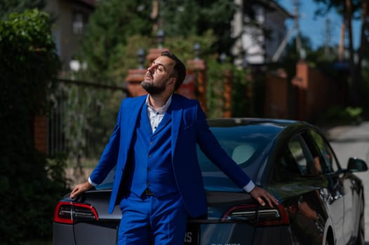 Caucasian bearded man in a blue suit stands near a black car in the countryside in summer