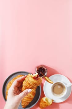 A woman's hand holds a croissant over a plate with fresh puff croissants, coffee and berry jam on a pink background. copy space.
