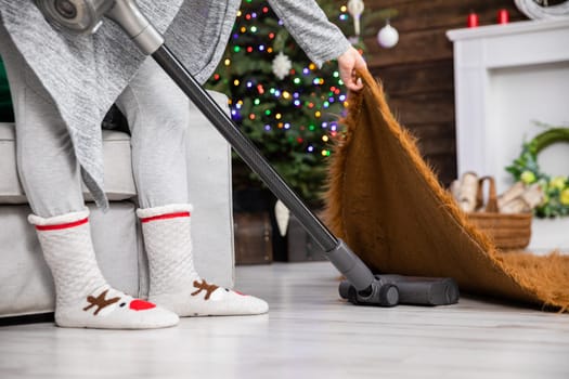 A woman lifts a rug up by the edge and vacuums thoroughly under it. The apartment decked out in decorations for reliving Christmas.