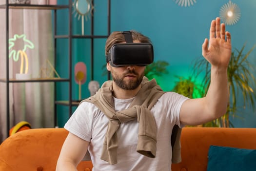 Excited man using virtual reality futuristic technology VR app headset helmet to play simulation 3D 360 video game watching film movie. Portrait of guy in goggles at modern home apartment room on sofa