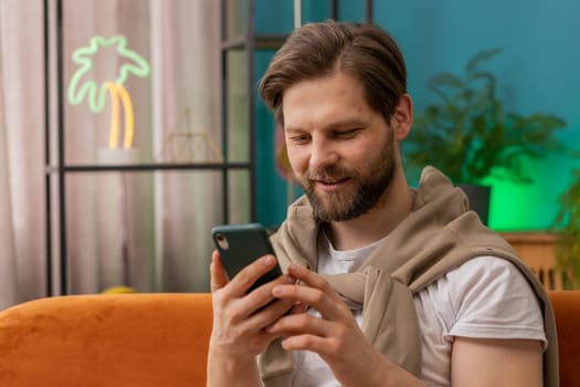 Caucasian man sitting on couch uses mobile phone smiles at home living room apartment. Young guy texting share messages content on smartphone social media applications online, watching relax movie