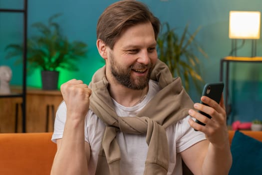 Happy man use mobile smartphone typing browsing say Wow yes found out great big win good news celebrate lottery jackpot doing winner gesture. Caucasian young guy at home in room sitting on orange sofa