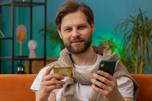 Smiling man using credit bank card and smartphone while transferring money, purchases online shopping cashless order food delivery at home apartment indoors. Happy young guy sitting in room on couch