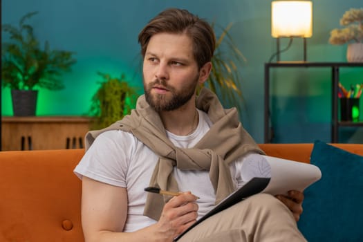 Caucasian man freelancer work with documents on home couch, reading information, business papers prepare financial report on laptop. Young guy accountant analyzing graph documents, calculate taxes