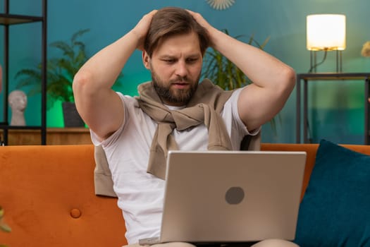 Displeased Caucasian man use laptop notebook typing browsing working, loses becoming surprised sudden lottery results bad news fortune loss fail computer virus. Young guy at home in room on couch