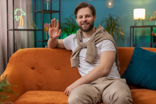 Hello. Caucasian handsome man smiling friendly at camera and waving hands gesturing hello, hi, greeting or goodbye, welcoming with hospitable expression at home. Guy sitting on sofa in living room