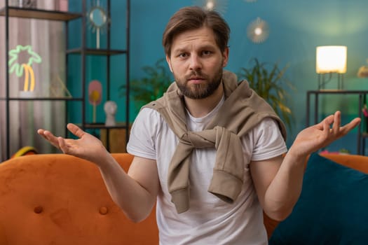 What. Why. Caucasian man raising hands in indignant expression asking reason of failure demonstrating disbelief irritation by troubles at home apartment. Young guy in living room sitting on sofa