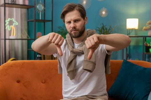 Dislike. Upset young man showing thumbs down sign gesture, expressing discontent, disapproval dissatisfied bad work at modern home apartment indoors. Displeased Caucasian guy in living room on sofa