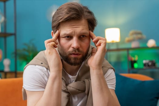 Displeased man rubbing temples to cure headache problem, suffering from tension and migraine, stress grimacing in pain high blood pressure at home room apartment. Caucasian guy on couch. Lifestyles