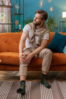 Young man suffer from painful severe knee joint ache, leg muscles, could not walk. Portrait of Caucasian guy massaging his knees illness cramps rheumatism at home room on sofa. Health care problem
