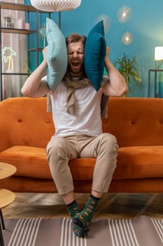 Repair work at neighbours. Irritated young man relaxing on couch cover ears with pillows annoyed by noisy neighbors suffer from headache wish silence. Thin walls at home flat without sound insulation
