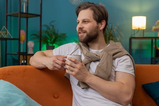 Portrait of smiling bearded man drinking a cup of warm coffee or herbal tea in the morning. Caucasian guy enjoying comfortable relaxing taking a break alone at home apartment living room on sofa