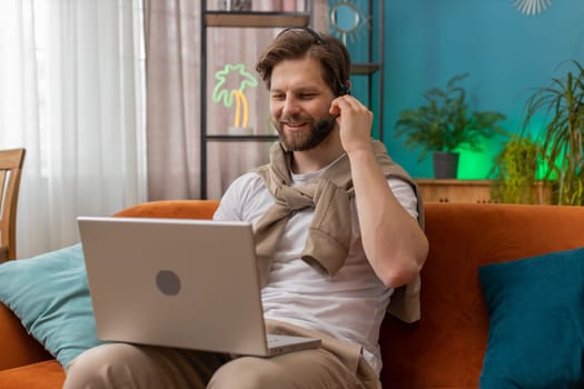 Caucasian man wearing headset, freelance worker, call center or support service operator helpline, having talk with client or colleague communication support waving hello. Guy at home room on sofa