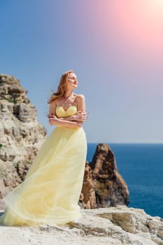 Woman yellow dress sea. Side view Young beautiful sensual woman in yellow long dress posing on a rock high above the sea at sunset. Girl in nature against the blue sky.