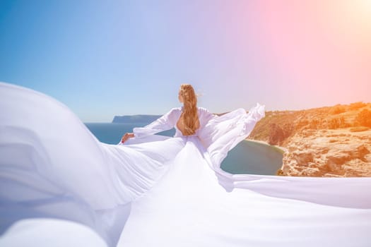 woman sea white dress. Blonde with long hair on a sunny seashore in a white flowing dress, rear view, silk fabric waving in the wind. Against the backdrop of the blue sky and mountains on the seashore