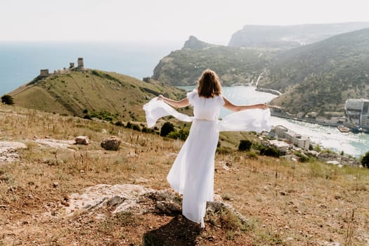 Happy woman in a white dress and hat stands on a rocky cliff above the sea, with the beautiful silhouette of hills in thick fog in the background