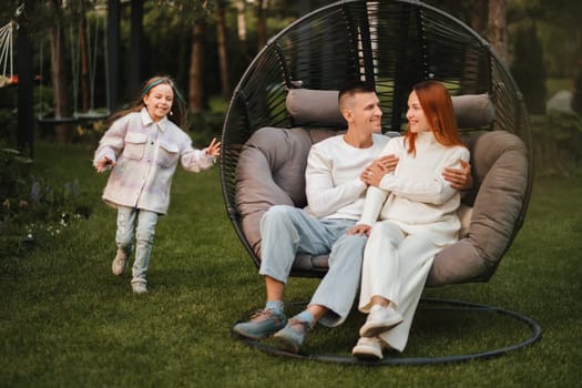 A happy family is sitting in a hammock on the lawn near the house.