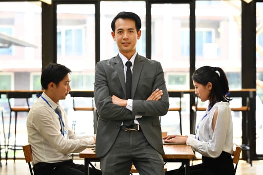 Portrait of confident asian male executive in suit standing with crossed arms and his colleagues sitting on background.