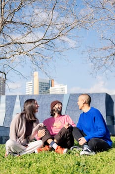 three young friends laughing happy sitting in the lawn at the city park in a sunny day, concept of friendship and urban lifestyle, copy space for text