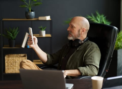 Smart and tech-savvy mid-aged man sits in chair at desk, engrossed in screen of phone. With bald head, silver beard, and air of handsomeness, multitasks, balancing phone, laptop, and cup of coffee. High quality photo