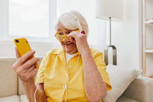 Happy elderly woman watching video call on phone and listening to music on headphones, surprise and open mouth, bright modern interior, lifestyle online communication. High quality photo