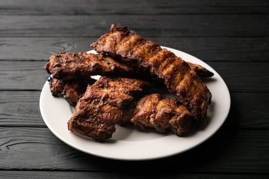 Freshly cooked pork ribs on a white round plate on a black wooden table in the background. Close-up, 45 degree angle view