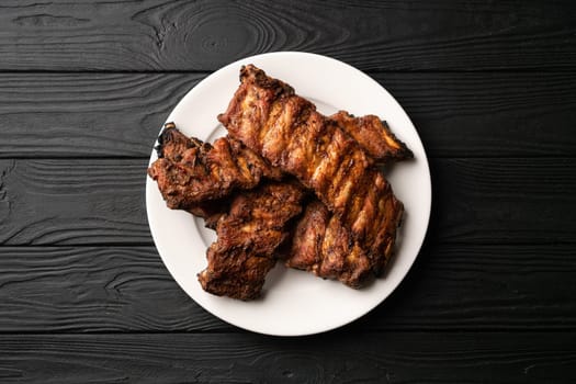 Freshly cooked pork ribs on a white round plate on a black wooden table in the background. Close up, top view