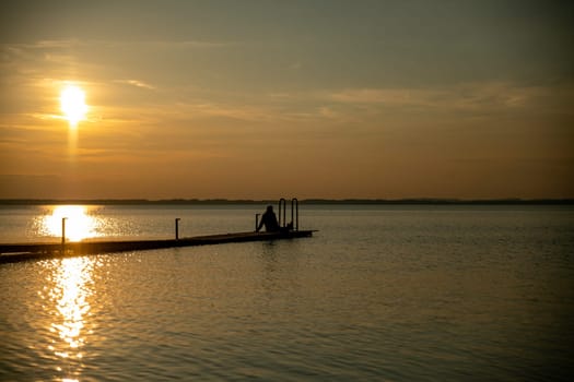 a woman sits on a wooden platform on the water during sunset with the horizon in the background