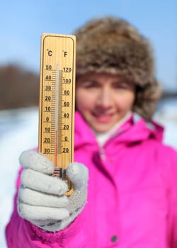 Young woman in pink jacket holds wooden thermometer outside on sunny but cold day, illustrating weather with freezing temperatures as low as -8 degrees Celsius.