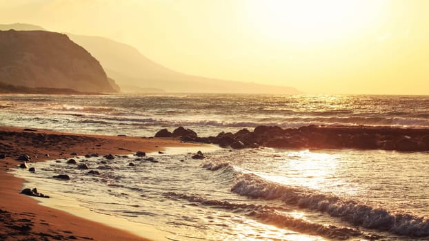 Empty wild beach lit by golden sunset. Small waves over rock, hills in hazy background.