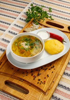 Chicken soup with vegetables in a white bowl served with mamaliga, red chilly pepper and sour cream.