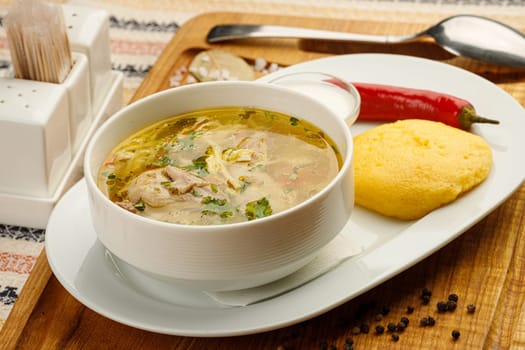 Chicken soup with vegetables in a white bowl served with mamaliga, red chilly pepper and sour cream.