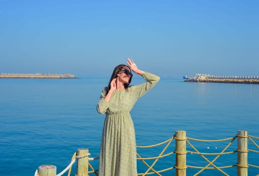 A young beautiful woman in a long dress stands alone on a pier by the sea, looking at the sun