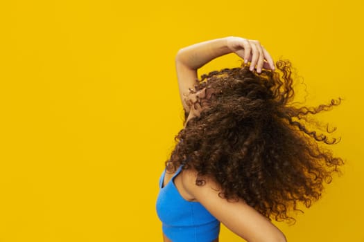 Woman with curly afro hair in a blue T-shirt on a yellow background dancing flying hair with sunglasses yellow, hand signs, look into the camera, smile with teeth and happiness, copy space. High quality photo