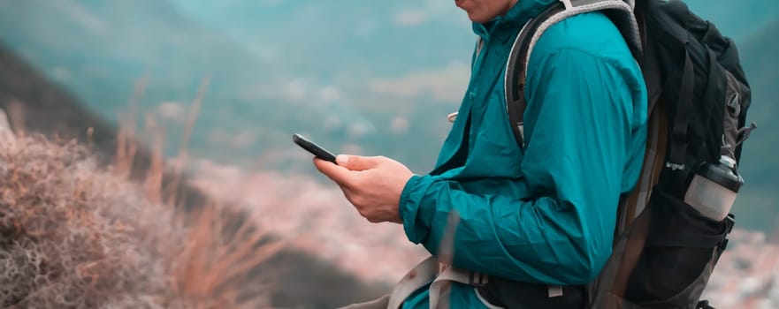 Young man in a turquoise hiking jacket holds a mobile phone in his hands, the beautiful mountains are on the background, a traveler with a backpack walks and uses downloaded maps on his smartphone.