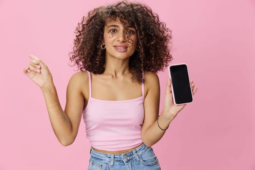 Woman blogger holding phone video call takes selfies, with curly hair in pink smile t-shirt and jeans poses on pink background, copy space, technology and social media, online. High quality photo