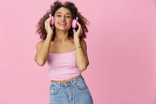 Happy woman wearing headphones with curly hair listening to music and dancing in a pink T-shirt and jeans on a pink background, copy space. High quality photo