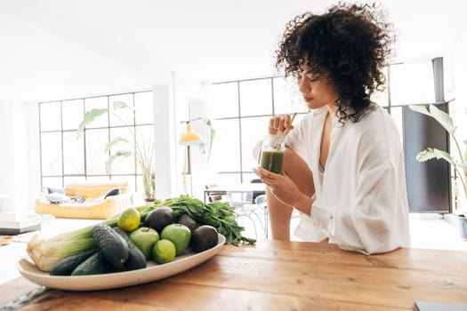 Young african american pretty woman relaxing in kitchen while having a green juice with bamboo straw in the morning. Home concept. Healthy lifestyle concept. Copy space.