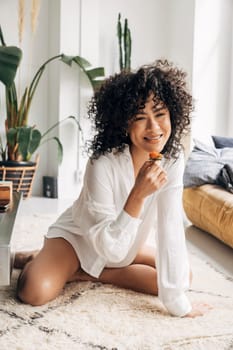 Young african american woman sitting on floor looking at camera eating strawberry in bright living room. Lifestyle.